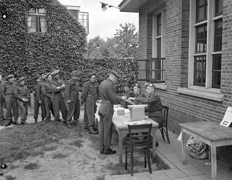 Black and white photo of members of the Canadian military waiting in line to vote at a polling station outside a brick building