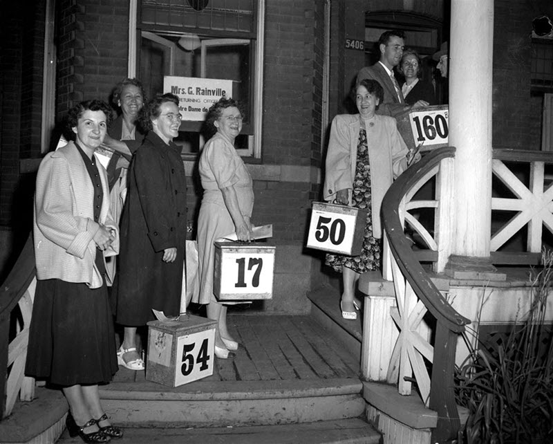 Black and white photo of a group, mostly women, waiting with metal ballot boxes on the steps of an election office