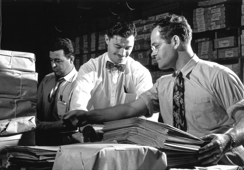 Black and white photo of three election workers organizing packages and documents on a table in a supply room