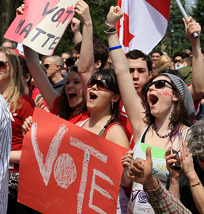 A crowd of young people outside cheer and pump their hands in the air. A young man waves a Canadian flag while a woman in front of him carries a handmade sign with the word 'vote' on it