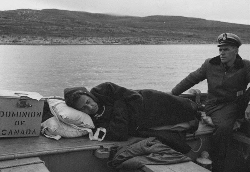 Black and white photo of an election worker lying on a bench on a boat with his eyes closed and a metal ballot box next to his head