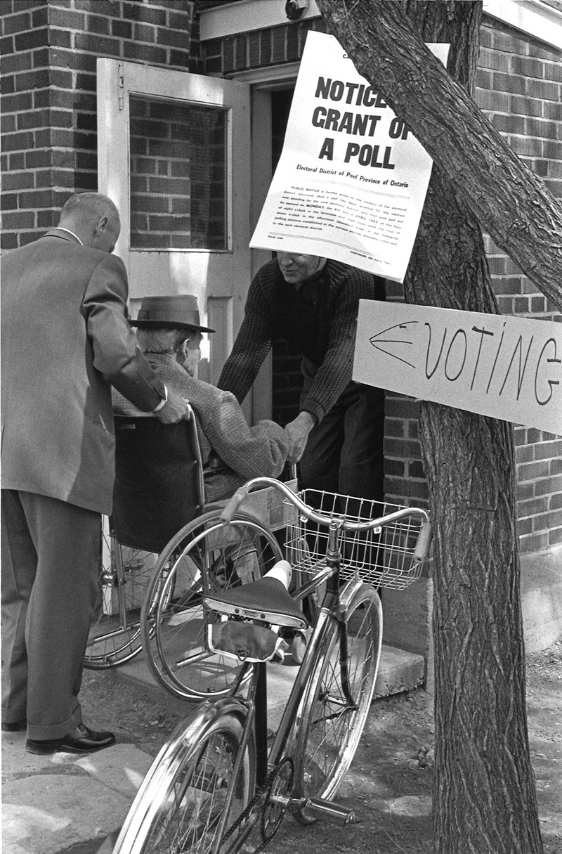 Photo en noir et blanc de deux hommes qui soulèvent un homme en fauteuil roulant au-dessus du seuil d'un bureau de scrutin.