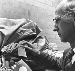 Photo en noir et blanc d'un homme d'âge moyen portant des lunettes qui tient dans ses mains deux étiquettes rectangulaires rattachées à de gros sacs de toile.