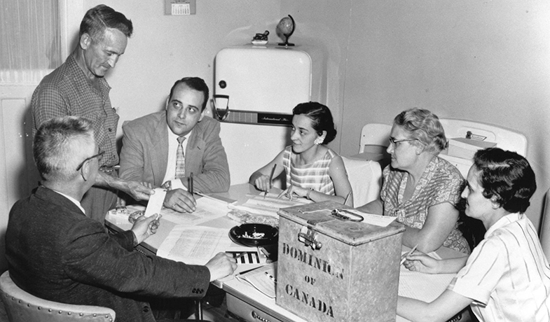 Black and white image of three men and three women sitting around a kitchen table piled with documents, an ash tray and metal ballot box.