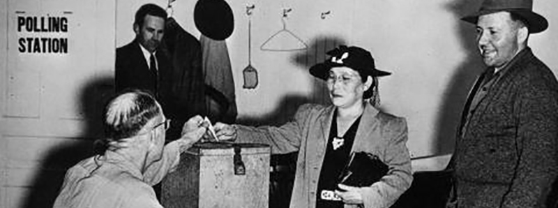 Black and white photo of a Japanese woman wearing a hat and long coat putting a ballot into a metal ballot box.
