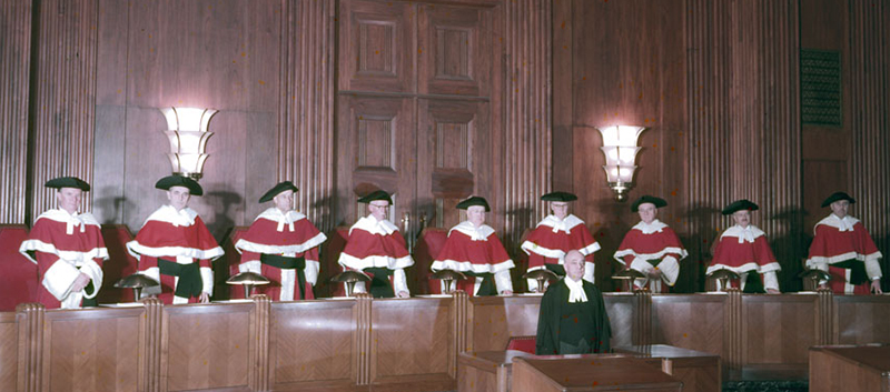 Neuf hommes portant des toges écarlates de la Cour suprême du Canada ornées de fourrure blanche sont debout derrière un long podium de bois