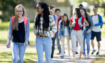 Two teenage girls in jeans walk along a paved with each other. Other young people wearing backpacks follow behind them