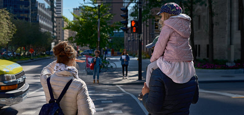 Une femme et un homme portant une petite fille sur ses épaules attendent à un passage pour piétons dans une rue de la ville