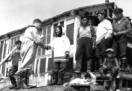 Black-and-white photo of an election worker and Inuit man shaking hands while several other Inuit men stand on the steps of a building watching.