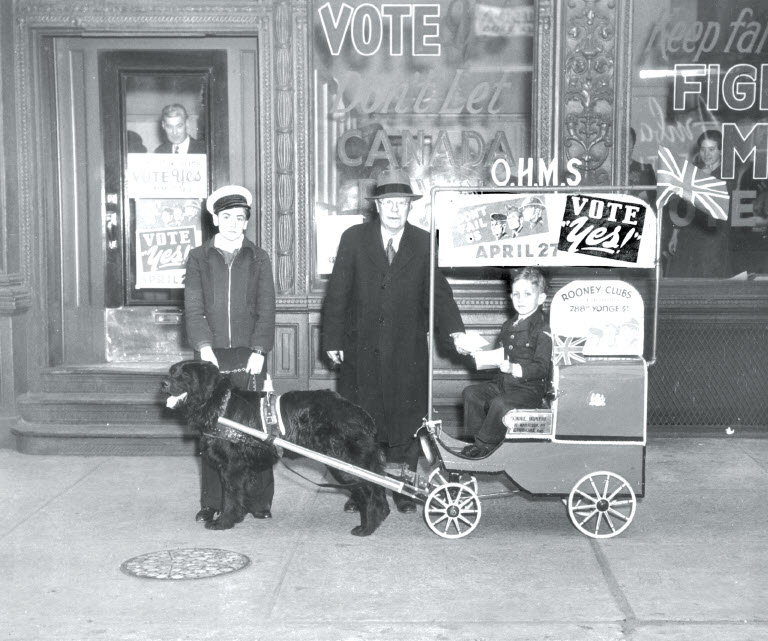 Photo en noir et blanc d'un enfant dans une charrette à chien utilisée pour promouvoir le « oui » au plébiscite de 1942 sur la conscription militaire. Un autre enfant et un homme accompagnent l'enfant.