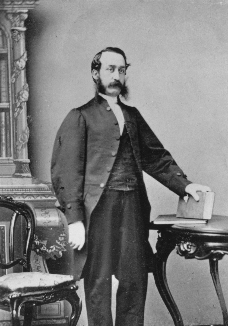 Photo en noir et blanc d'Édouard J. Langevin vêtu d'une redingote noire, posant avec un livre pour un portrait officiel.