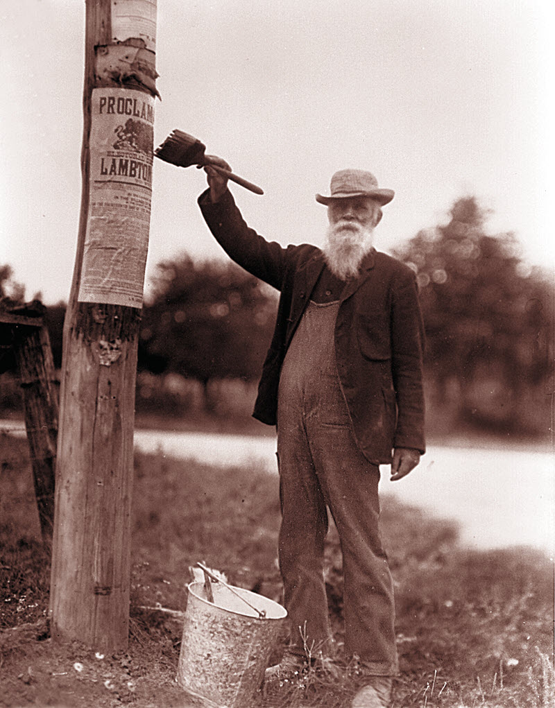 Photo d'un homme âgé qui colle une affiche annonçant une élection sur un poteau au moyen d'un pinceau et d'un seau de colle.