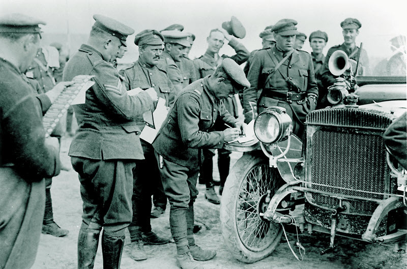 Black and white photo of a dozen soldiers in First World War uniforms holding and marking ballots as they crowd around an antique car.