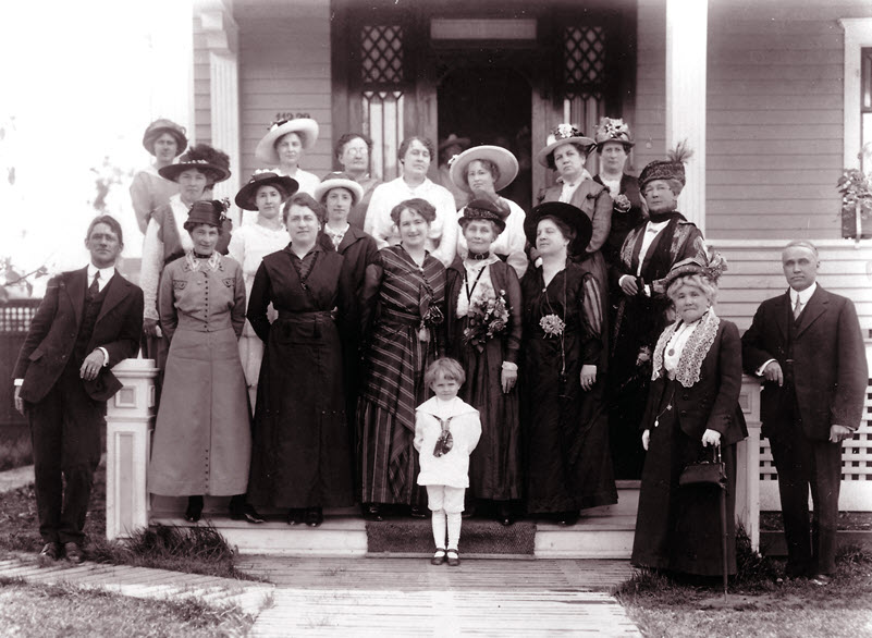 Black and white photo of a group of mostly women in long dresses and hats standing on the porch of a home.