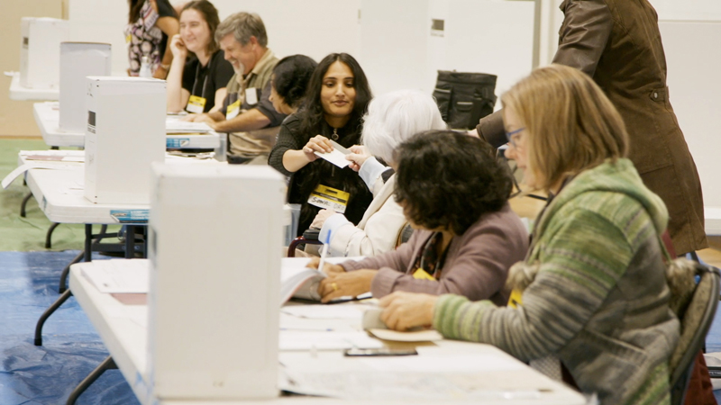 Photo d'un bureau de scrutin montrant des préposés au scrutin assis à une longue table sur laquelle se trouvent des urnes.