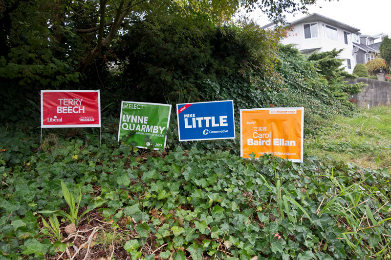 Photo des pancartes électorales de quatre candidats soutenus par quatre partis différents, placées dans l'herbe.