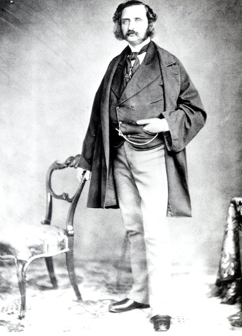 Black-and-white photo of Alexandre-Édouard Kierzkowskiman standing in a dark vest and frock coat and light-coloured trousers with one arm resting on the back of a chair.